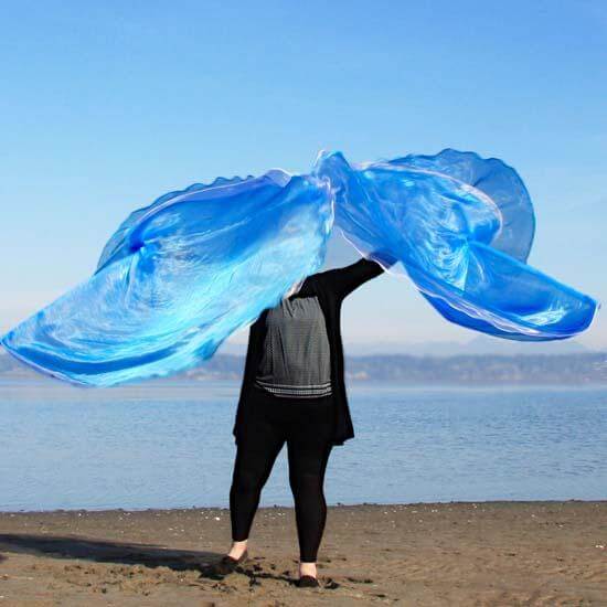 Front view of a woman worshipping with large sheer blue and white worship flags on a beach, flags extended wide in both arms with ocean and mountains in the background.