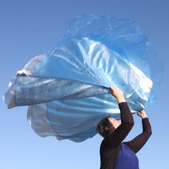 Woman worshipping outdoors with large flowing blue and silver worship flags against a clear sky, expressing movement and praise.