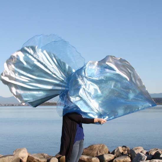 Worshipper standing on rocky shoreline waving large blue and silver worship flags, capturing fluid motion against a peaceful lake and mountain backdrop.