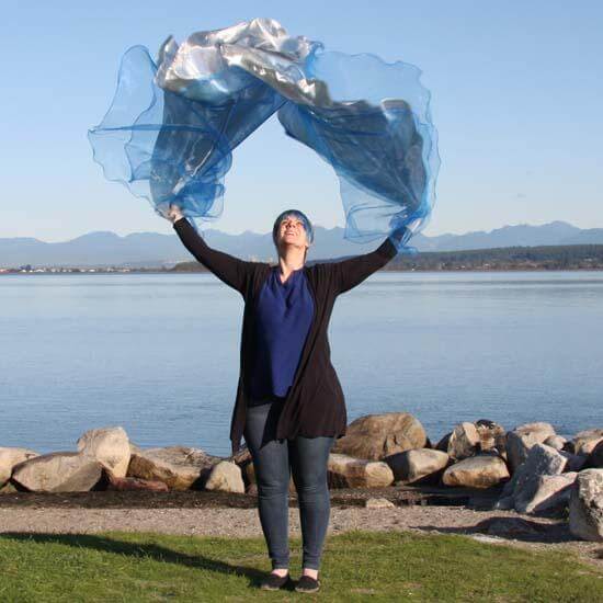 Woman worshipping with flowing blue and silver flags by the waterfront, arms raised in praise under a clear sky with mountains in the background.