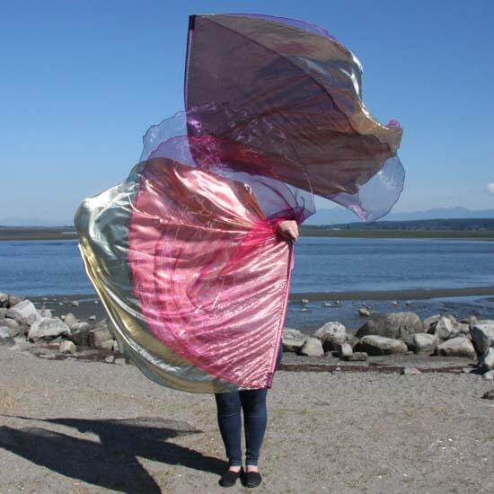 Worshiper standing on a beach holding layered worship flags in shimmering gold, scarlet, and purple, flowing in the wind with ocean and sky in the background.