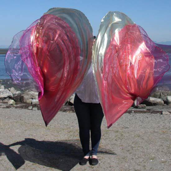 Worshipper holding large layered worship flags in purple, gold, and scarlet, shimmering with iridescent light at a beachside setting, representing royal praise and biblical symbolism.
