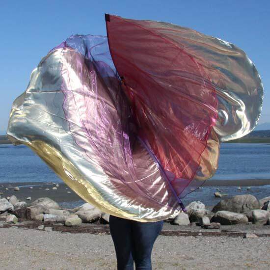 Worshiper at the shoreline holding layered worship flags in gold, purple, and scarlet, billowing in the wind against a backdrop of rocks, water, and blue sky.