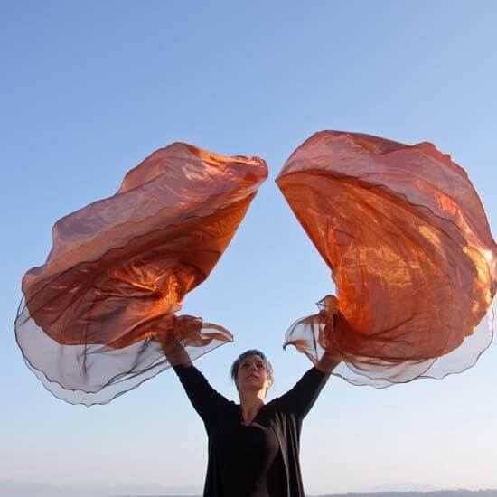 Woman worshipping outdoors with a pair of copper and bronze shimmer worship flags raised high against a clear blue sky