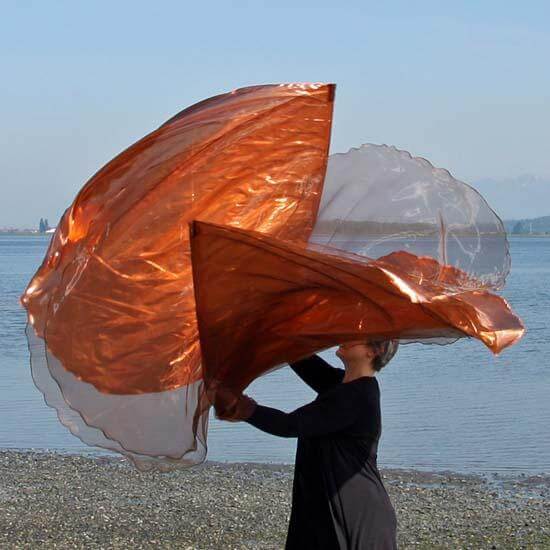 Worshipper by the water holding large, flowing worship flags in bronze and copper shades with sheer, iridescent shimmer against a clear sky.