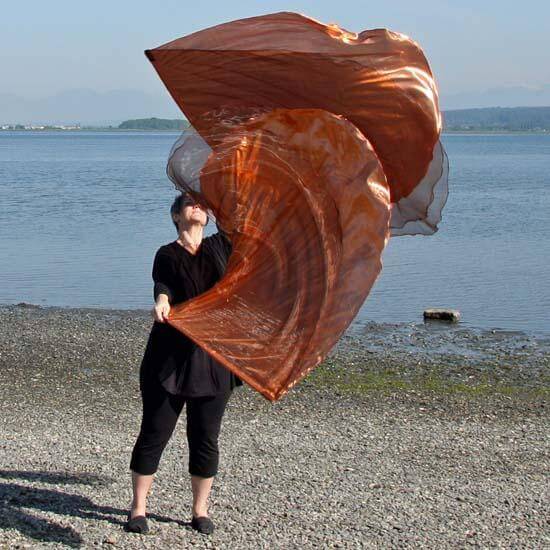 Woman worshipping with large bronze and copper worship flags near a lakeshore, with flags billowing upward against a calm blue sky and water backdrop.