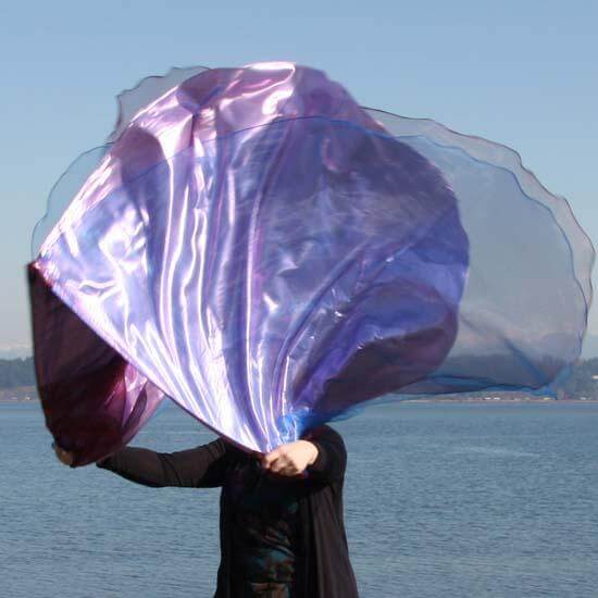 "Worship flags in shades of pink, blue, and wine captured mid-movement near the shoreline, showcasing layered iridescent fabric and expressive motion.