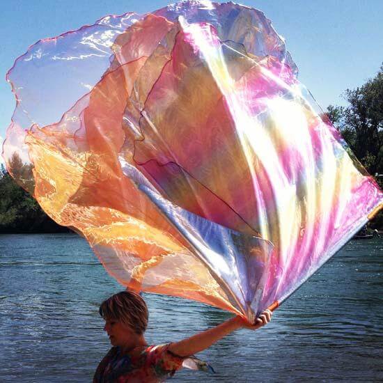 Dancing with flowing handmade worship flags, shimmering in orange and rainbow colors by the river.