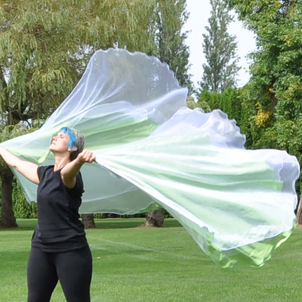 A woman with short blue hair and black clothing stretches her arms wide, holding large white and green translucent worship flags that billow dramatically behind her. She stands on a vibrant green lawn surrounded by tall trees and lush foliage, creating a serene and worshipful outdoor atmosphere.