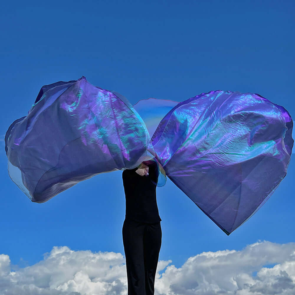 A person in black worships with a pair of flowing, iridescent purple and rainbow flags raised high against a deep blue sky. The luminous fabric reflects green and blue tones, creating a celestial effect. Puffy white clouds float in the background, emphasizing the heavenly atmosphere of the worship moment.