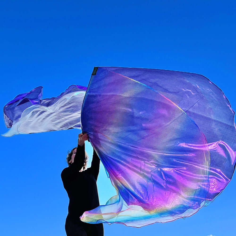 A worshipper dressed in black waves vibrant worship flags under a bright blue sky. The flags shimmer with rainbow hues of purple, lilac, and subtle gold, creating a radiant arc of light. Their movement forms a dynamic swirl that visually lifts the atmosphere in a celebration of praise and beauty.