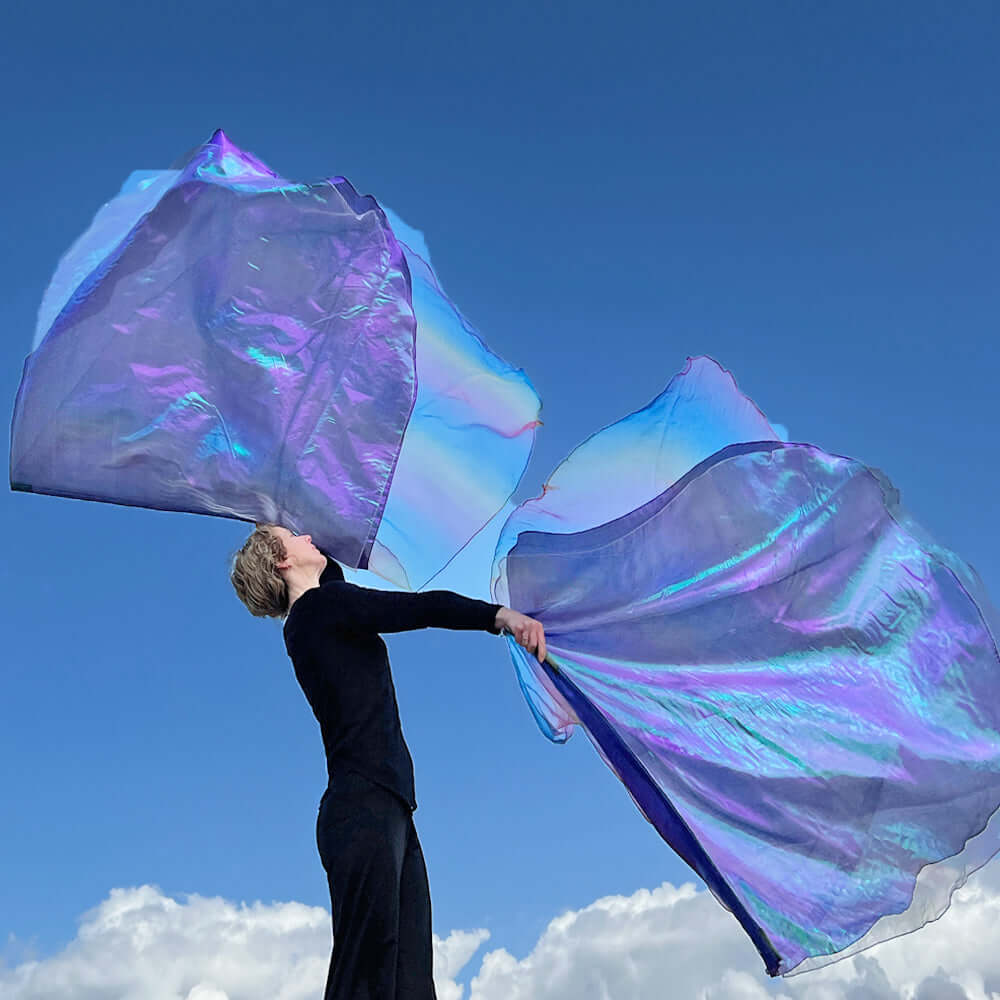A woman dressed in black waves two iridescent worship flags in the air against a vivid blue sky. The flags shimmer with layers of purple, blue, and hints of rainbow, catching the light with a heavenly glow. She stands on a raised surface with white clouds below, creating a powerful image of exaltation and spiritual elevation.
