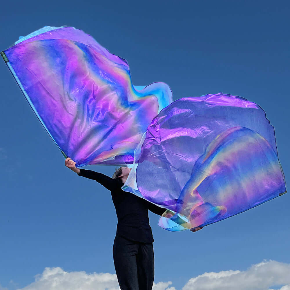 A dancer dressed in black lifts large worship flags against a vivid blue sky. The flags shimmer with radiant shades of purple, lilac, and rainbow iridescence, catching the sunlight and creating a vibrant, heavenly swirl above her. The movement appears joyful and powerful, evoking the beauty of expressive worship.