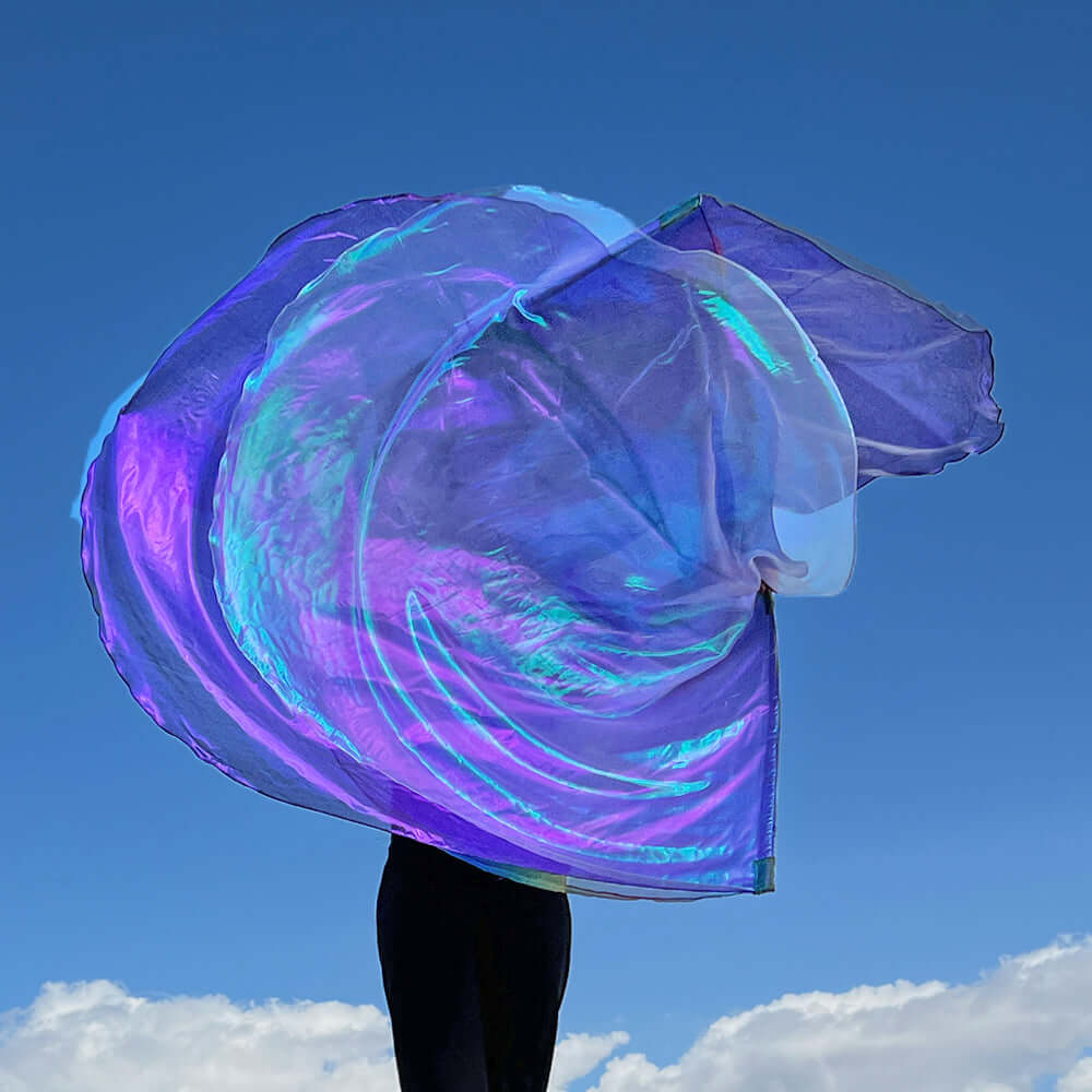 A pair of iridescent worship flags swirl together mid-air against a deep blue sky. The flags, in shades of purple, lilac, and crystal, shimmer with rainbow reflections as they arc in unison. The flagger, dressed in black, is mostly hidden behind the flowing movement, making the fabric appear like angelic wings unfolding above the clouds.
