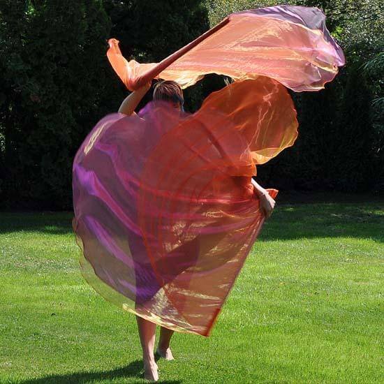 A barefoot person dances gracefully on green grass, holding large worship flags in motion. The flags are sheer and flowing in shades of orange, scarlet, and hints of magenta, creating a vibrant swirl against a backdrop of lush greenery. The movement and color convey energy and expressive worship.