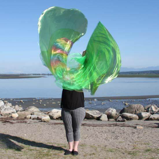A person in checkered pants and a black top raises vibrant green worship flags that shimmer with hints of rainbow colors. The translucent flags create a dynamic swirl against a serene beach and mountain backdrop under a clear blue sky.