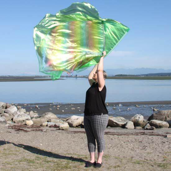 A worshipper in black and checkered pants lifts a large green worship flag that shimmers with rainbow hues in the sunlight. The translucent flag flows above a beach shoreline, framed by rocks and distant mountains under a clear sky.