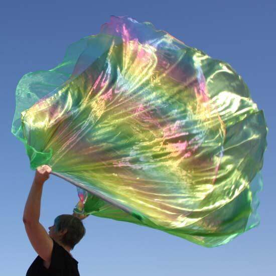 A worship flag dancer waves a large, iridescent flag that shimmers in green and rainbow hues against a clear blue sky. The flag has flowing, layered fabric with reflective highlights in yellow, pink, and blue, creating a vibrant, radiant effect. The person is dressed in black, with arms raised, expressing a dynamic moment of worship.
