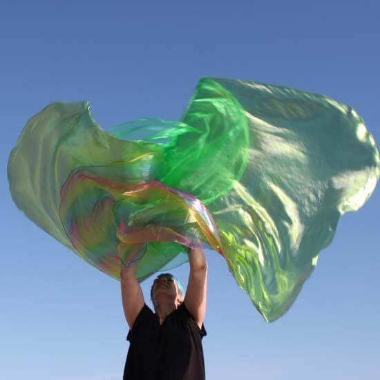 A worshipper in black lifts a pair of large green worship flags that shimmer with hints of rainbow iridescence. The translucent fabric catches the light as it flows upward, glowing against a soft blue sky in a powerful moment of expressive praise.