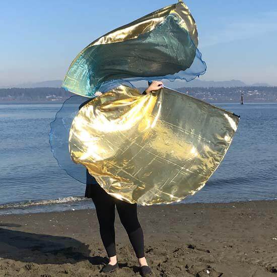 A person stands on a sandy beach holding up two large metallic worship flags. The flags shine with gold and blue tones, catching the sunlight. The ocean and distant hills are visible in the background under a soft blue sky.