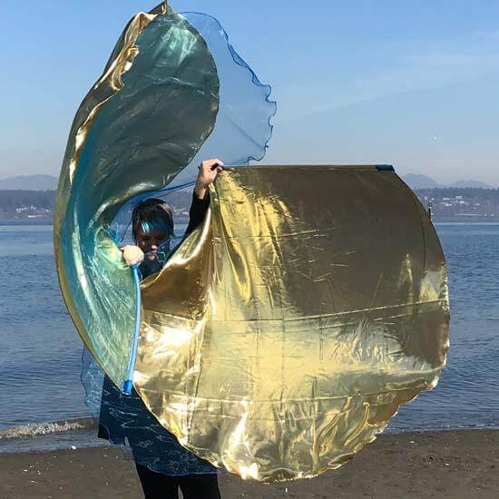 A person stands on a sandy beach holding up two large metallic worship flags. The flags shine with gold and blue tones, catching the sunlight. The ocean and distant hills are visible in the background under a soft blue sky.