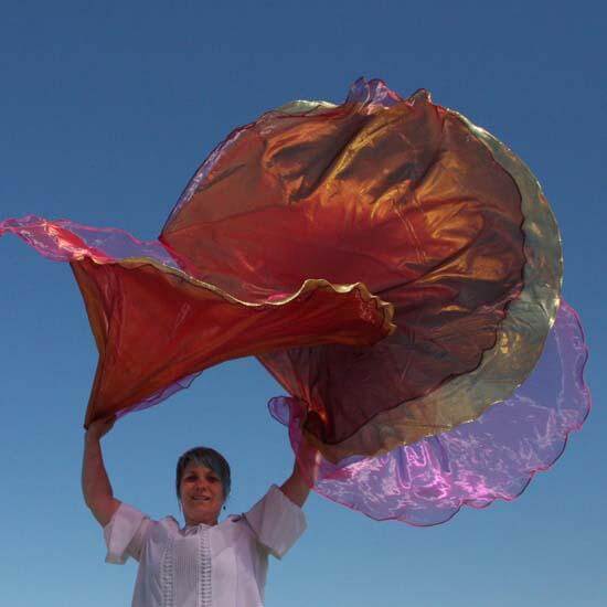 A woman waves layered worship flags against a vivid blue sky. The flags swirl in rich hues of coral, gold, and wine with a sheer pink layer catching the sunlight, creating a vibrant, radiant motion in the air.