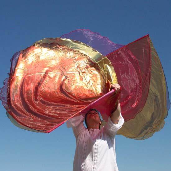 A worshipper lifts a pair of worship flags against a clear blue sky. The flags feature vibrant coral, shimmering gold, and deep wine colors, creating layered waves of fabric that catch the sunlight. The person wears a white top and stands in a posture of praise, adding a dynamic sense of movement and devotion to the image.