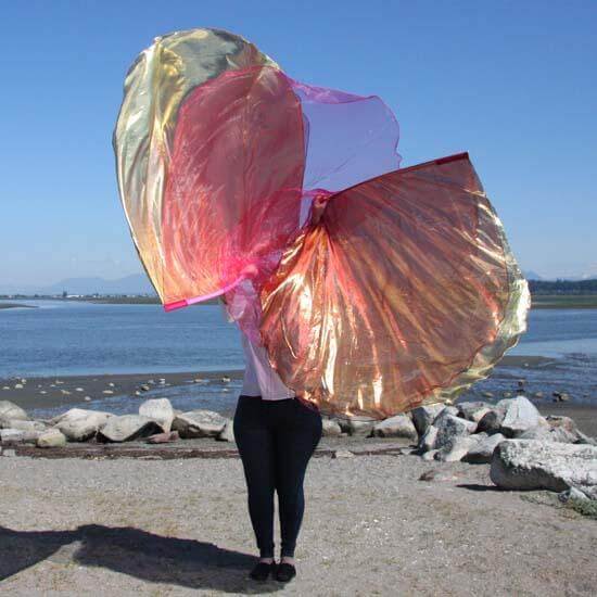 A person stands on a rocky beach holding a pair of layered worship flags in front of them. The flags shimmer in hues of coral, gold, and wine with a sheer pink layer on top, glowing under the bright sunlight. The ocean and mountains stretch out in the background under a clear blue sky.