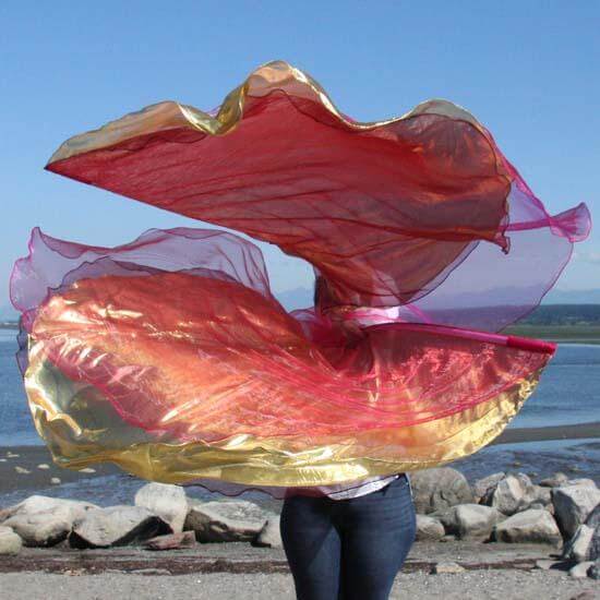 A person waves a layered set of worship flags in a stunning outdoor setting near the water. The flags shimmer in vibrant coral, gold, and wine tones, with a translucent pink layer adding depth and movement to the swirling fabric.