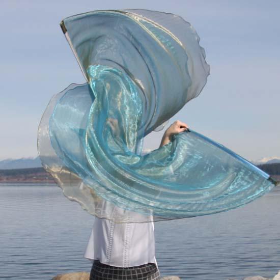A person stands in front of a calm lake, holding up shimmering worship flags in soft olive green and light blue tones. The flags swirl in a graceful arc, reflecting light with a metallic sheen.