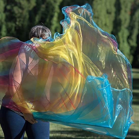 Woman worshipping with a flowing, rainbow-colored worship flag featuring vivid hues of yellow, blue, and red in a park with tall trees in the background.
