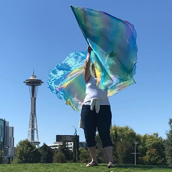 Woman waving iridescent rainbow-colored worship flags in a grassy park near the Space Needle in Seattle, Washington, under a clear blue sky.