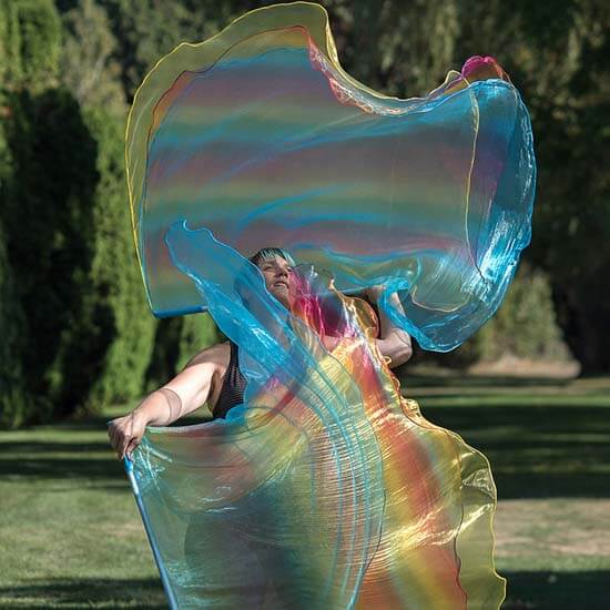 Woman worshipping with two large, multicolor worship flags outdoors on a sunny day, featuring flowing fabric in rainbow hues of blue, yellow, pink, and green.