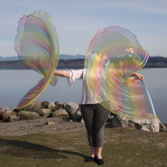 Person standing by a calm lake holding two iridescent worship flags in yellow and rainbow tones, with mountains and soft sky in the background.