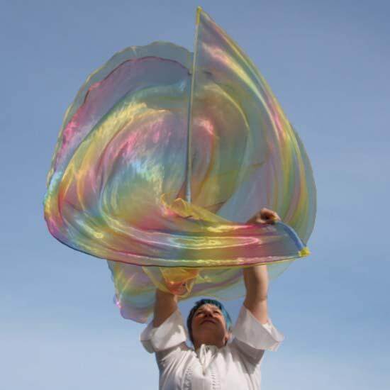 Woman holding large worship flag in shimmering yellow and rainbow colors, billowing against a soft blue sky with a sense of movement and joy.