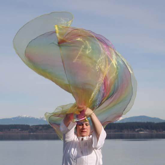Woman worshipping outdoors with flowing yellow and rainbow worship flags, raised overhead with graceful movement by a lake and distant mountain backdrop.