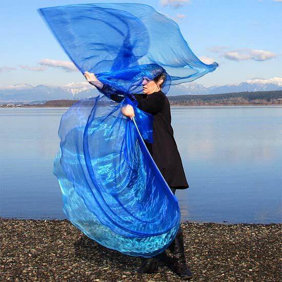 Woman worshipping at the water’s edge with vibrant blue worship flags swirling in motion, mountains and sky reflected on the calm lake behind her.