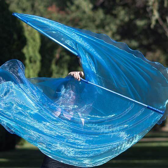 Worshipper holding a large blue flag mid-motion, creating a sweeping wave effect against a backdrop of trees in a sunlit park.