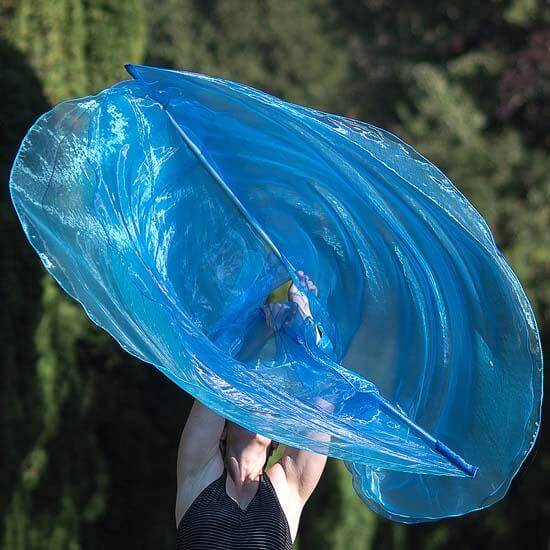 Person lifting a vibrant blue worship flag high overhead, creating a dynamic circular motion with translucent fabric against a forested background.