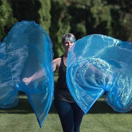 Woman worshipping outdoors with flowing blue worship flags in motion, set against a lush green background of trees and sunlight.