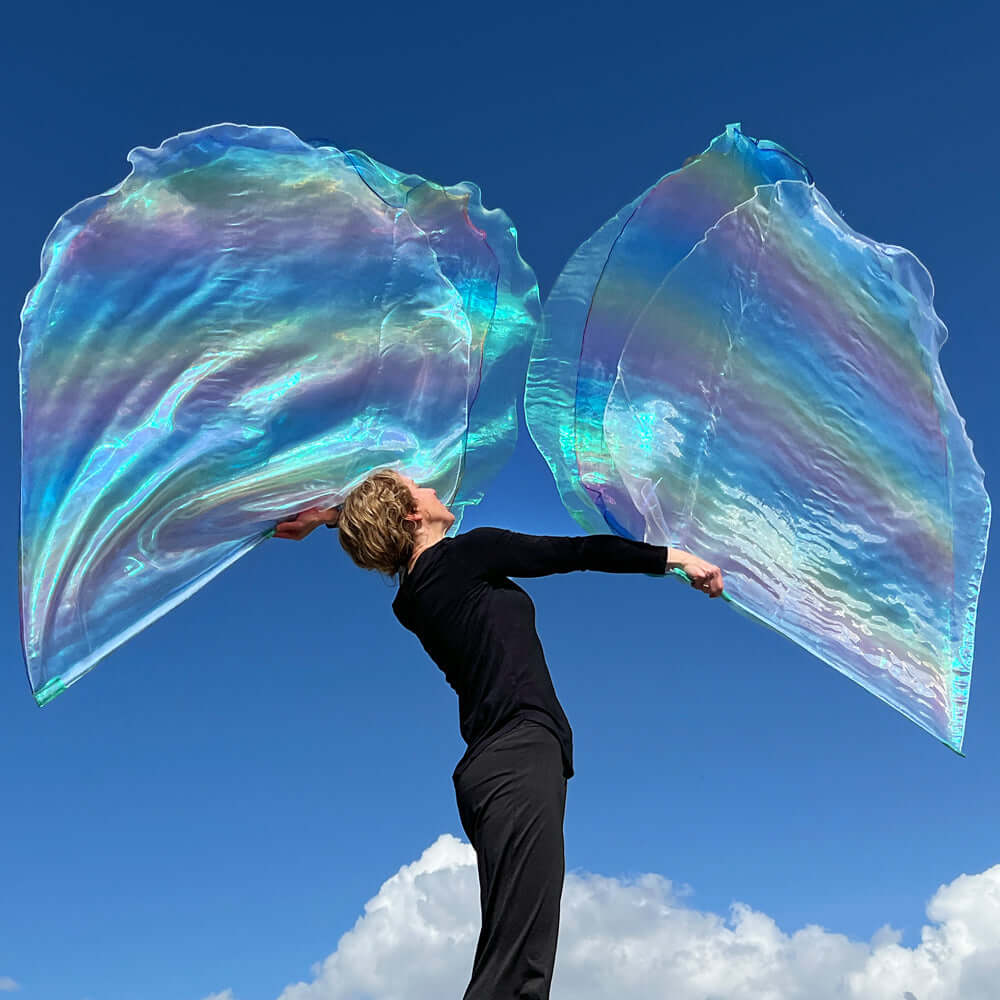 Woman worshipping outdoors with large, iridescent seaglass and rainbow crystal worship flags flowing upward against a deep blue sky and clouds.