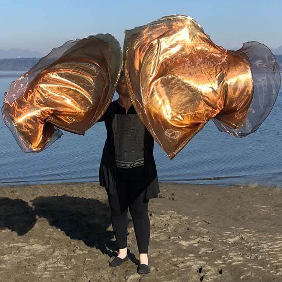 A woman stands on the sand holding bronze and gold worship flags mid-movement, with a calm waterfront in the background.