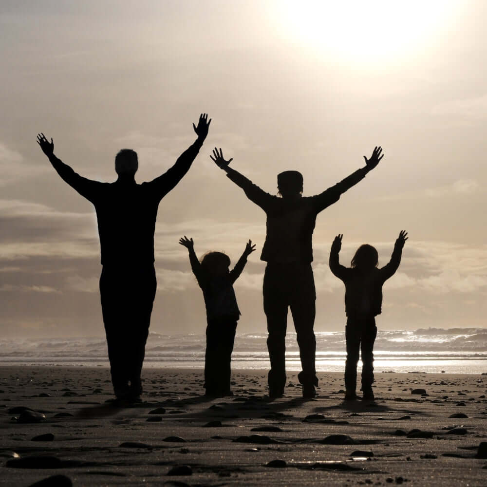 Silhouetted family lifting hands in worship at sunset on the beach, representing generational praise and raising children in faith.
