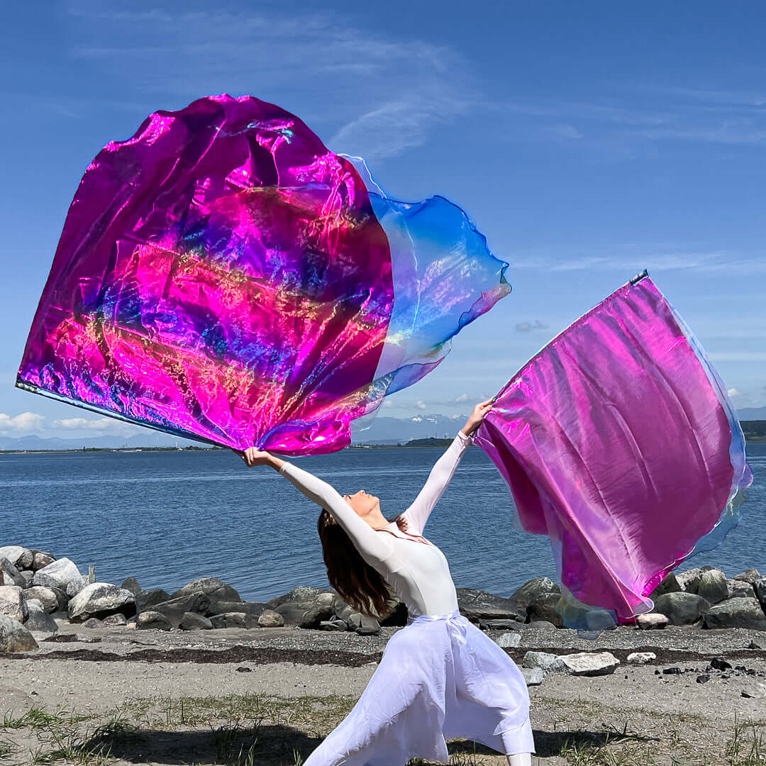 A worship dancer strikes a powerful pose at the edge of a rocky beach, arms outstretched as she waves two vibrant worship flags. The left flag bursts with pink, rainbow, and crystal hues, while the right flag is a solid shimmering pink. She is dressed in flowing white garments, and the bright sky and distant mountains create a majestic backdrop for her expressive movement.