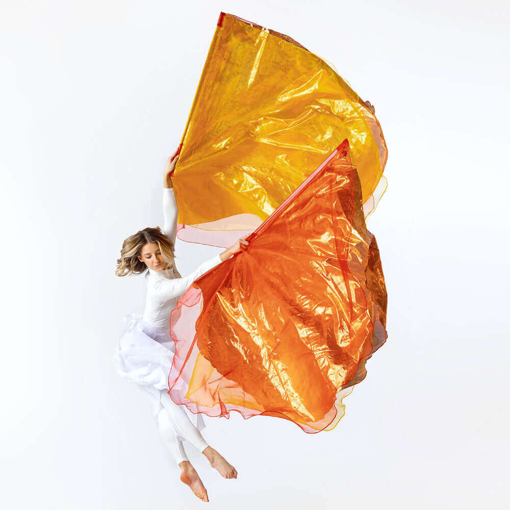 woman in white worship attire leaping in midair while holding large red copper and yellow worship flags, expressing dynamic praise and worship.