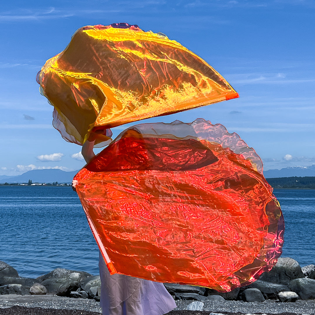  Two worship flags billow in the air against a coastal backdrop. The top flag glows with golden yellow and soft orange tones, while the lower flag radiates with intense fiery red and orange hues. The dancer, partially hidden by the motion, stands at the shoreline dressed in white, creating a vivid contrast with the vibrant flags and deep blue sky.