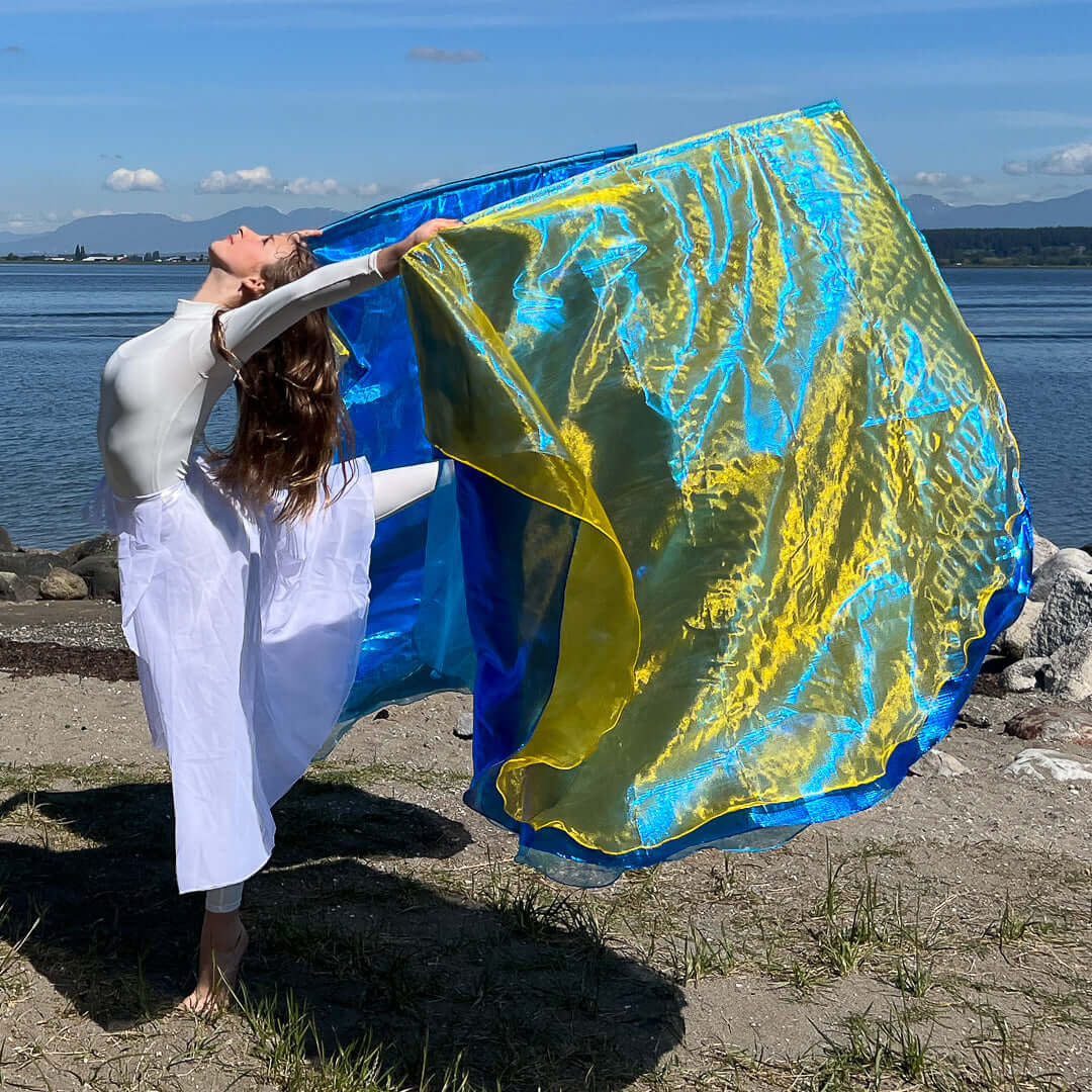 A worship dancer in a flowing white outfit lifts her arms in a graceful arch on the shoreline, holding vibrant worship flags in bright blue and radiant yellow. The flags ripple like waves in the wind, catching the sunlight as the ocean and distant mountains frame the moment of joyful praise.