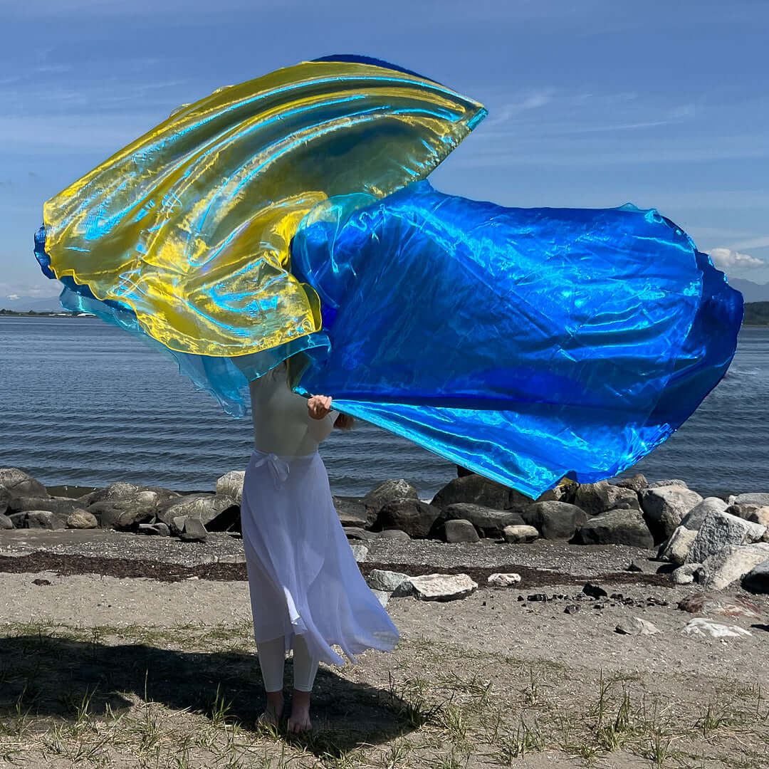A worshipper stands at the edge of a rocky shoreline, cloaked in white and lifting two brilliant worship flags—one vivid blue and the other a luminous yellow. The wind fills the translucent fabric, creating a stunning wave-like motion against the calm ocean and sky.