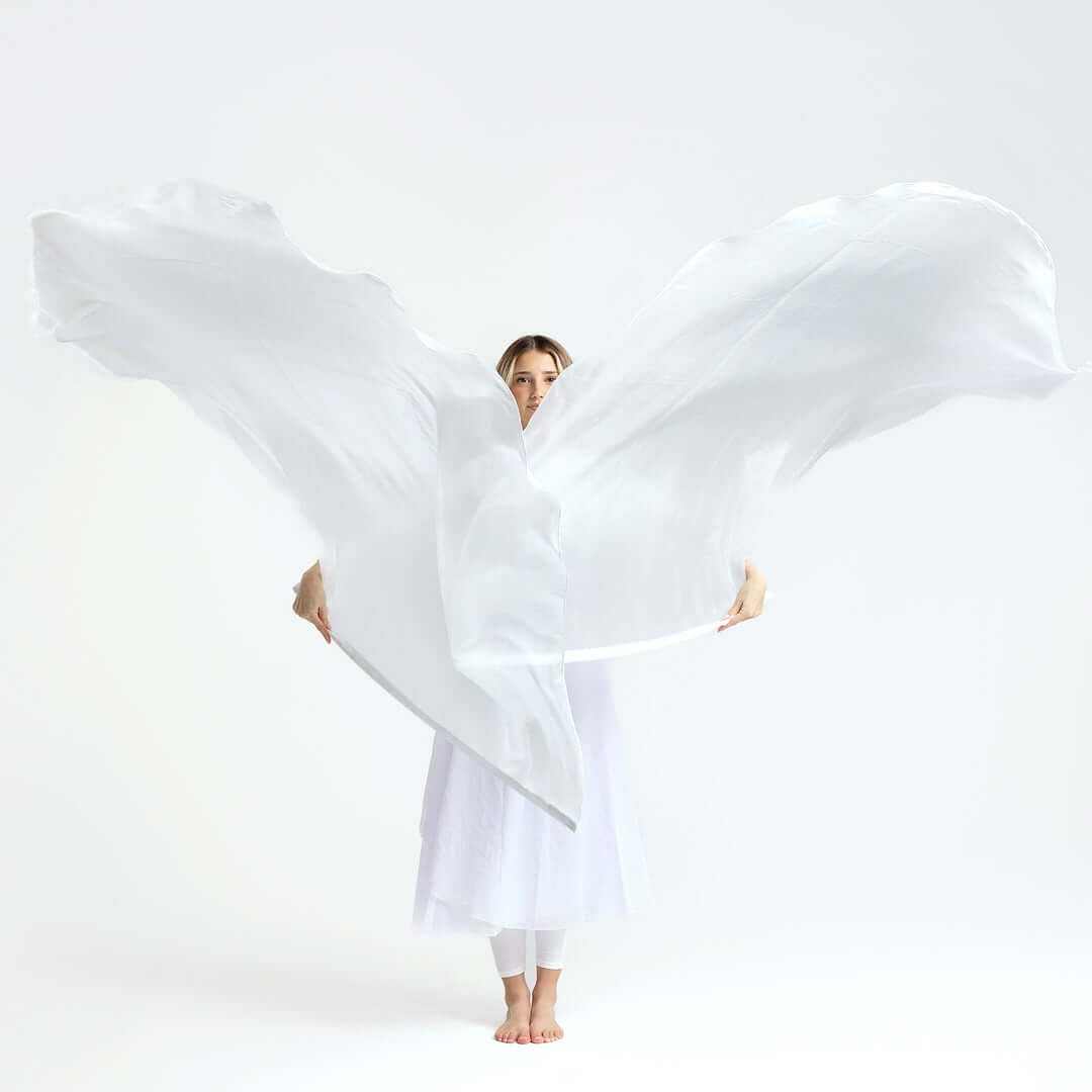 Woman in white praise attire standing barefoot with two large white worship flags spread wide, partially covering her in a powerful still worship pose.
