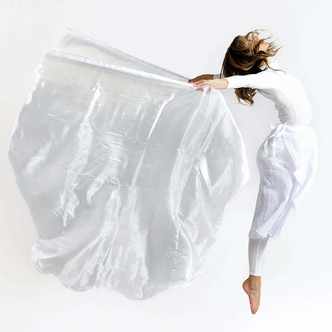 Woman in white worship attire captured mid-jump with a large flowing white flag, representing freedom, holiness, and expressive praise.